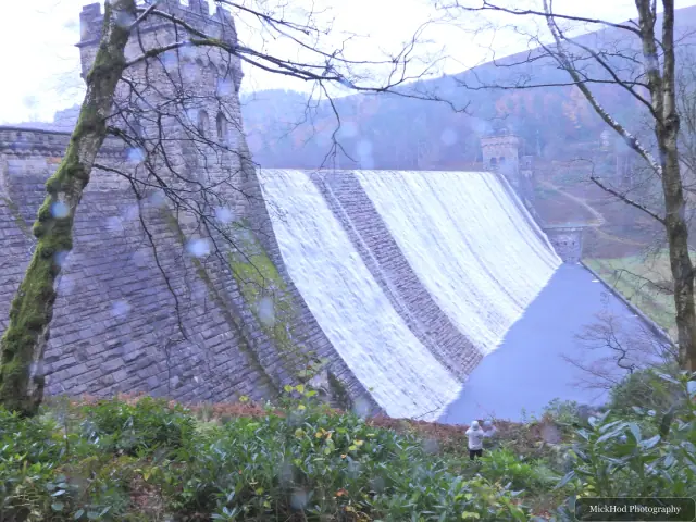The dam and reservoir are seen through the bare trees on the wooded hillside.