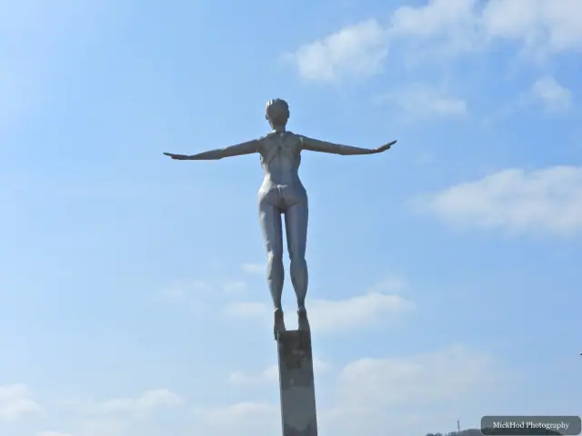 A tall bronze statue stands with outstretched arms against a bright, cloud-dappled sky.