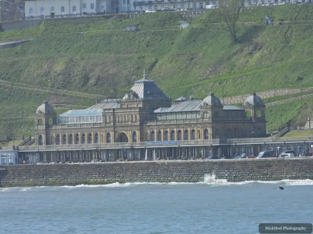 Scarborough Spa sits on the seafront below the green hillside, viewed across the blue water.