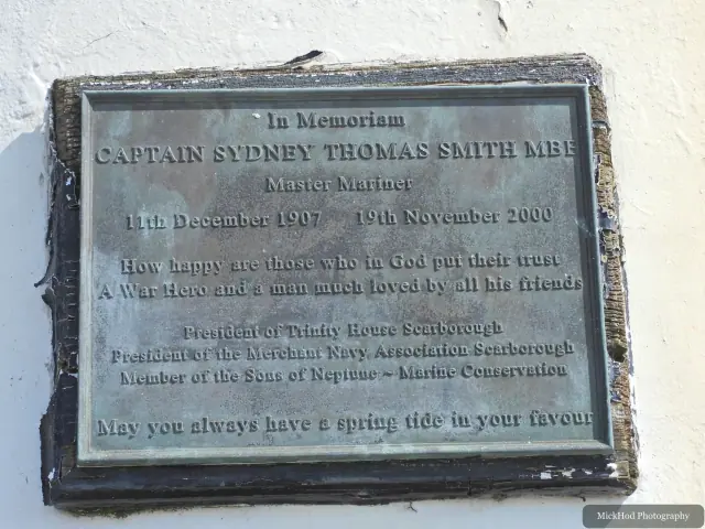 A memorial plaque honours Captain Sydney Thomas Smith MBE on a sunlit white wall.
