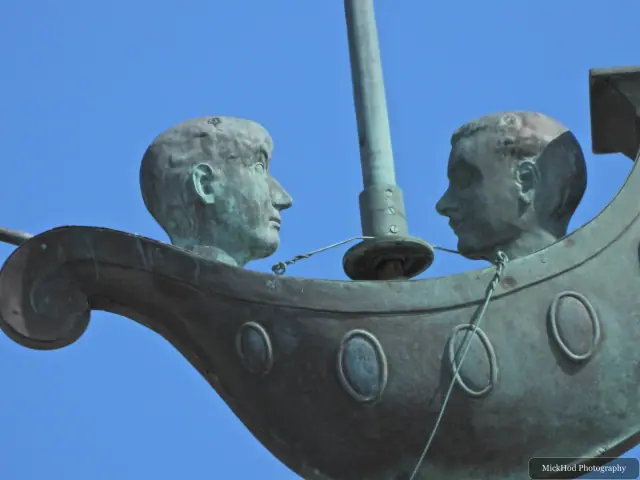 A close view of the bronze boat and its two seated figures, with the mast rising above them.
