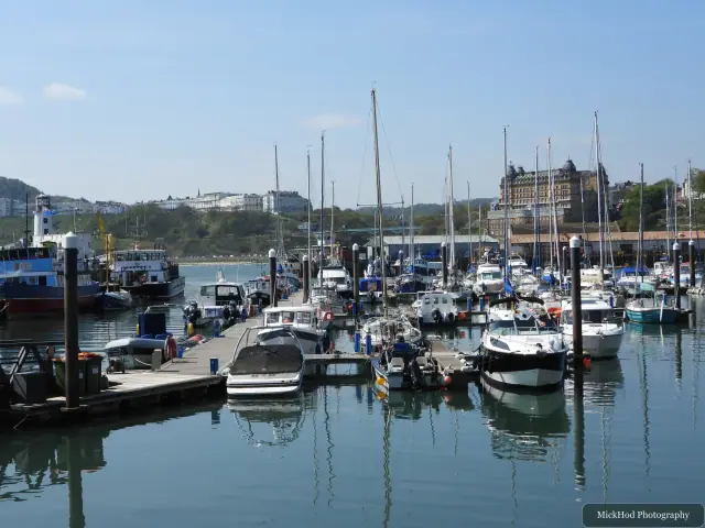Yachts and motorboats sit in the sunlit harbour, backed by the town and headland.