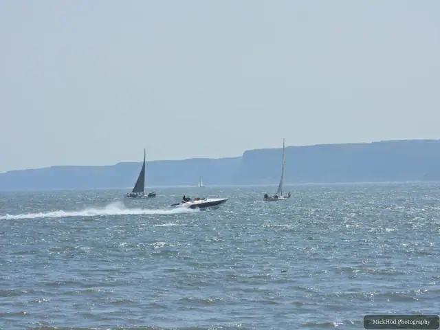 Several small boats sail offshore, with the distant headland stretching across the horizon.